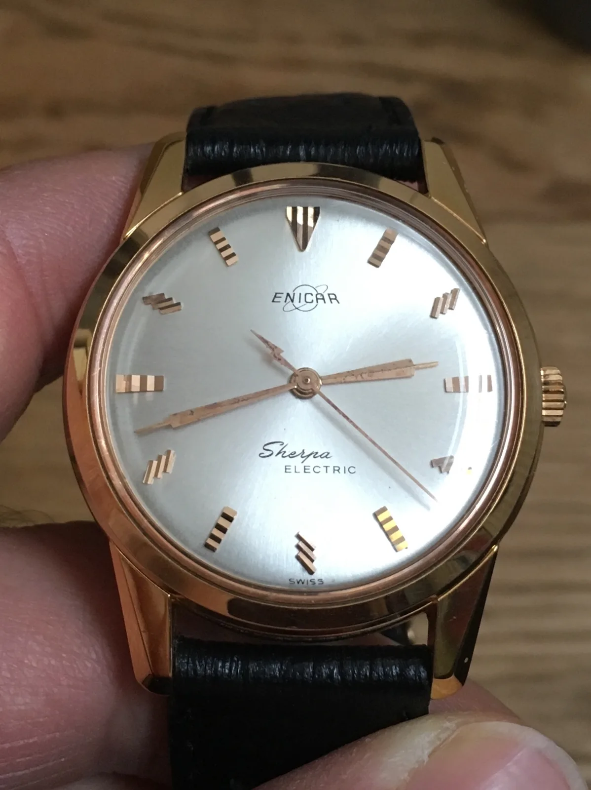 Gold-plated Sherpa Electric with a silver dial and gilt markers on a black strap, held in the hand