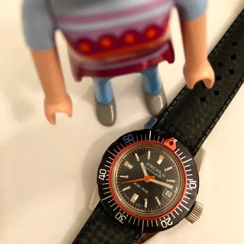 Sherpa Mini-Dive with a black dial, red pointer ring and outer bezel next to a toy figure