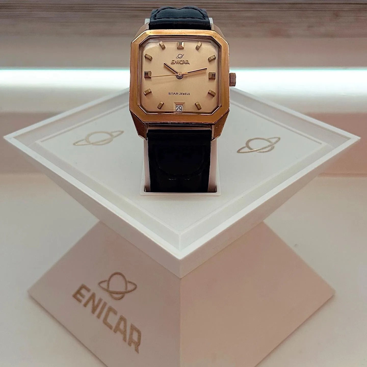 Gold-coloured Star Jewels with a square case on a black strap, on an Enicar display stand