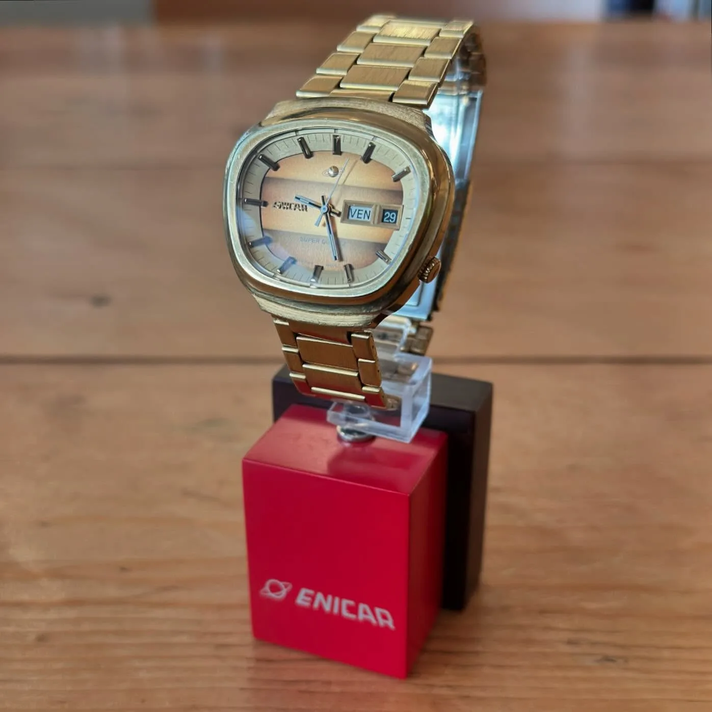 Gold-coloured Super Quartz in an asymmetric case on its bracelet, on an Enicar stand